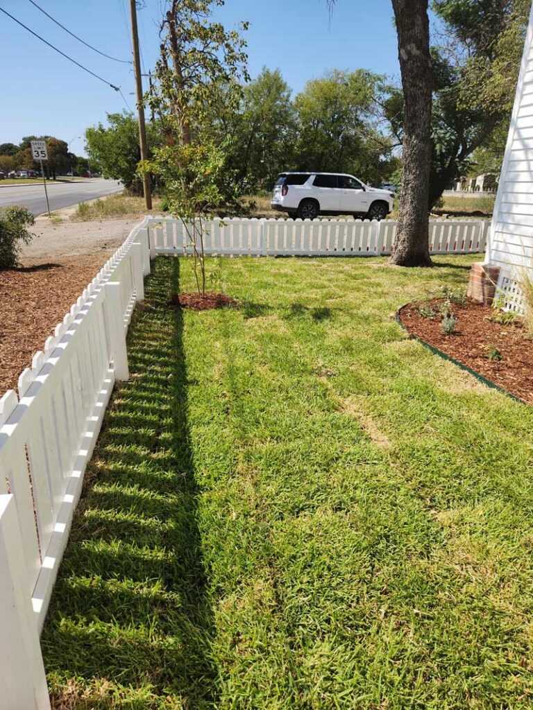Freshly installed sod lawn with a dark mulched flower bed along a picket fence, an example of sod and mulch installation by Trees Forever Nursery in Brownwood, TX